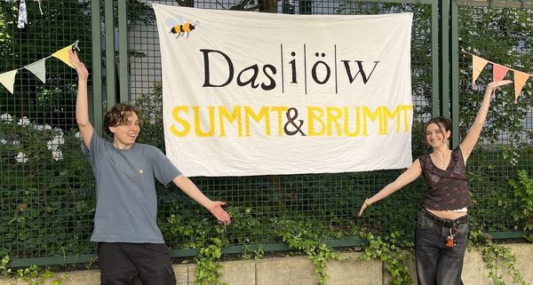 Zwei junge Menschen präsentieren ein Banner mit der Aufschrift "Das IÖW summt und brummt" in einem geschmückten Hinterhof.